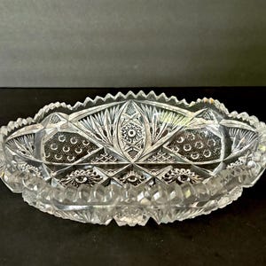 Puede incluir: Un cuenco de cristal transparente de forma ovalada con intrincados patrones de vidrio tallado. El cuenco presenta un borde festoneado y diseños geométricos y florales detallados. El cuenco de cristal es probablemente para servir o exhibir.