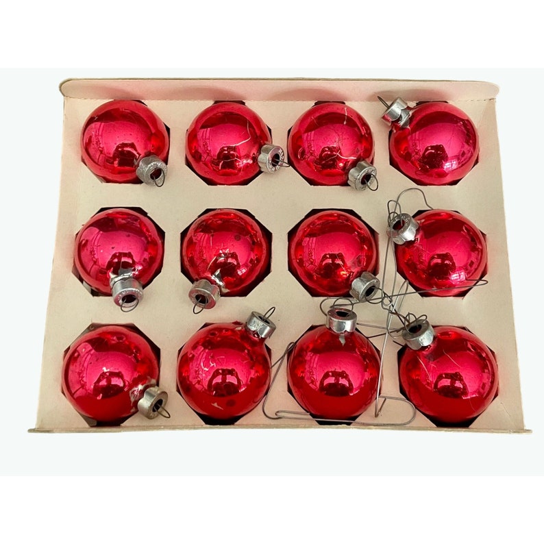 Puede incluir: Una caja de doce adornos navide&ntilde;os de vidrio rojo con tapas plateadas. Los adornos est&aacute;n dispuestos en un patr&oacute;n de cuadr&iacute;cula en la caja.