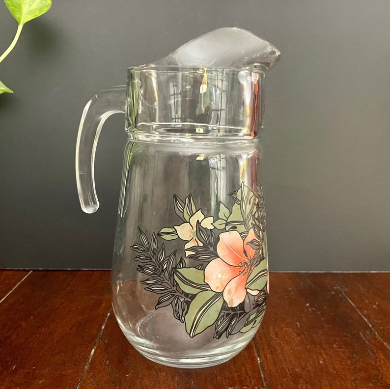 Puede incluir: Una jarra de vidrio transparente con un dise&ntilde;o floral. El dise&ntilde;o presenta flores rosas, blancas y verdes con contornos negros. La jarra tiene un asa y una tapa.