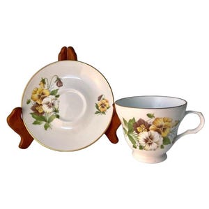 May include: A white teacup and saucer set with a floral pansy design in yellow, brown, and white. The teacup has a curved handle and a gold rim. The saucer is round with a gold rim. The set is displayed on a wooden stand.
