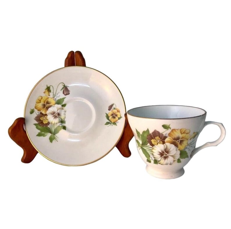 May include: A white teacup and saucer set with a floral pansy design in yellow, brown, and white. The teacup has a curved handle and a gold rim. The saucer is round with a gold rim. The set is displayed on a wooden stand.