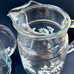 May include: Two clear glass pitchers with white floral designs and gold trim. The pitchers are empty and have a vintage look.