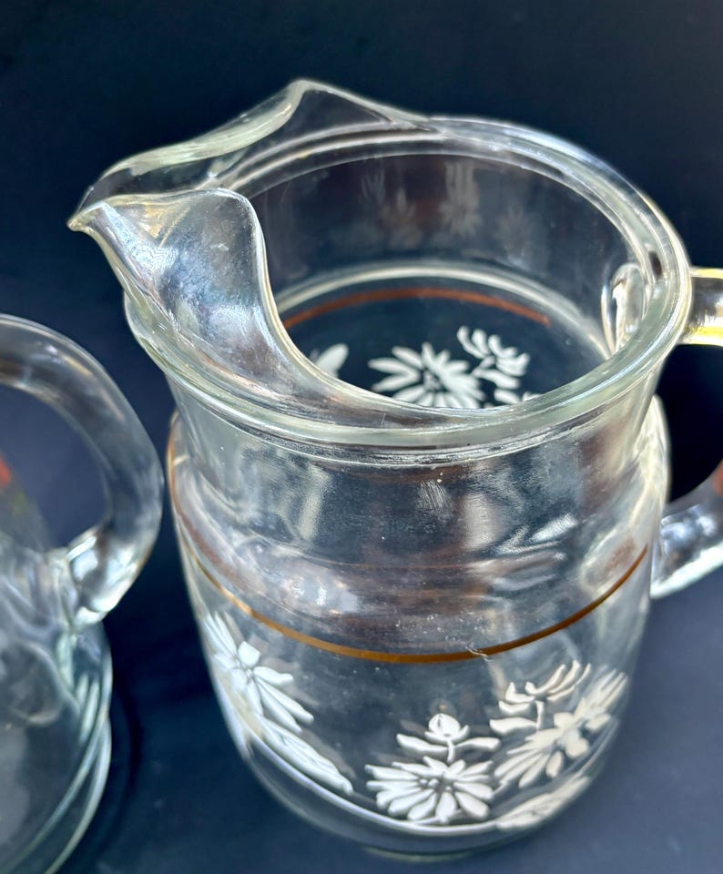 May include: Two clear glass pitchers with white floral designs and gold trim. The pitchers are empty and have a vintage look.