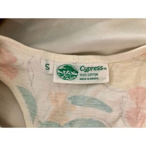 May include: Close-up of a small, off-white tank top with a floral pattern in pink, peach, and green. The tag reads "Cypress" and "100% Cotton Made in Brazil". The size is "S".