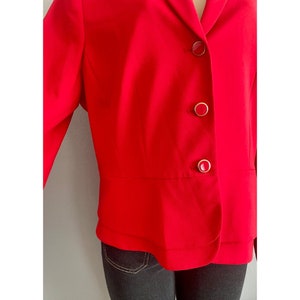 May include: A vibrant red blazer featuring a notched lapel and three circular buttons down the front. Each button has a red center surrounded by a thin gold-toned metallic rim. The jacket has a structured fit with a horizontal seam at the waist.