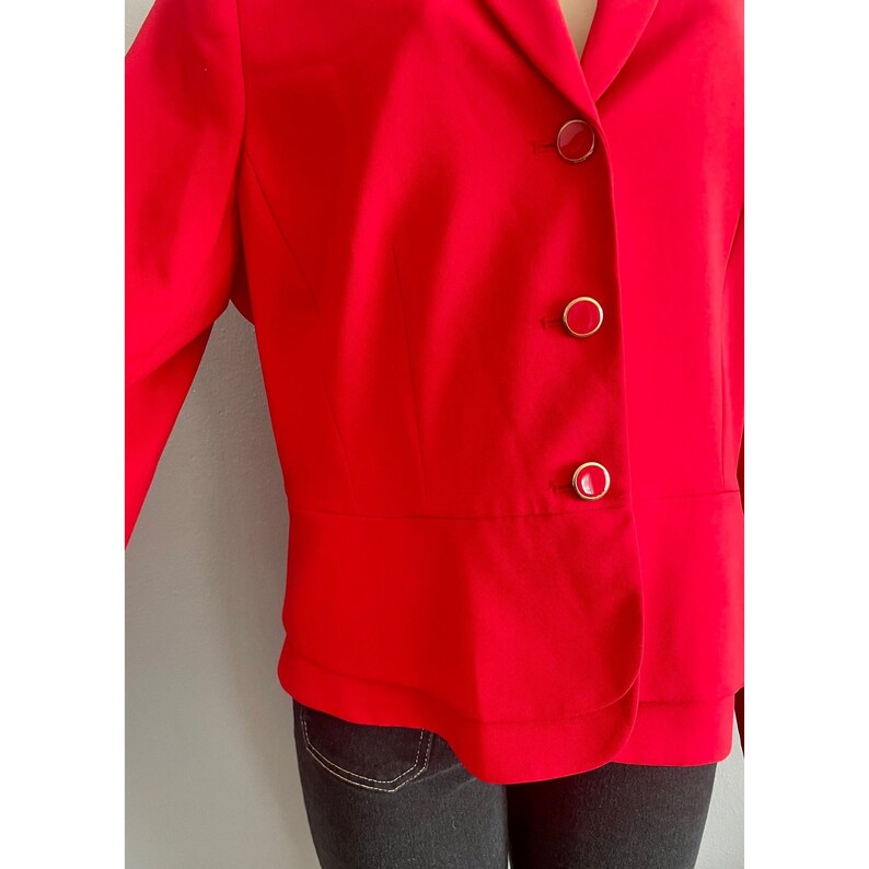 May include: A vibrant red blazer featuring a notched lapel and three circular buttons down the front. Each button has a red center surrounded by a thin gold-toned metallic rim. The jacket has a structured fit with a horizontal seam at the waist.