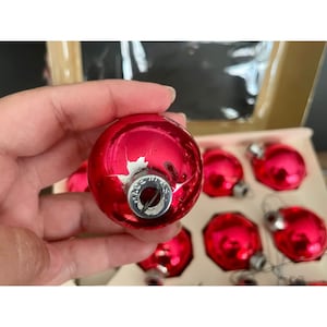 Puede incluir: Un conjunto de ocho adornos navide&ntilde;os de vidrio rojo con tapas plateadas. Los adornos est&aacute;n en una caja y est&aacute;n siendo sostenidos por una mano. Los adornos son todos del mismo tama&ntilde;o y forma. Los adornos tienen un acabado brillante.