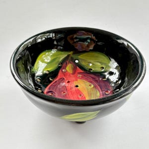 Puede incluir: Un colador de cerámica negro con un diseño de fruta pintado a mano. El interior presenta una fruta roja y morada con hojas verdes. El colador tiene agujeros de drenaje y se apoya en tres pequeños pies. El borde y el exterior son negros.