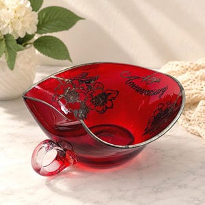 May include: A deep red glass bowl with a silver-colored rim and a small handle on one side. The bowl features a black floral design and the text 40th Anniversary printed in a cursive script on the interior surface of the glass vessel.