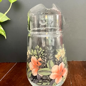 Puede incluir: Una jarra de vidrio transparente con un dise&ntilde;o floral. El dise&ntilde;o presenta flores rosas y amarillas con hojas verdes sobre un fondo negro.
