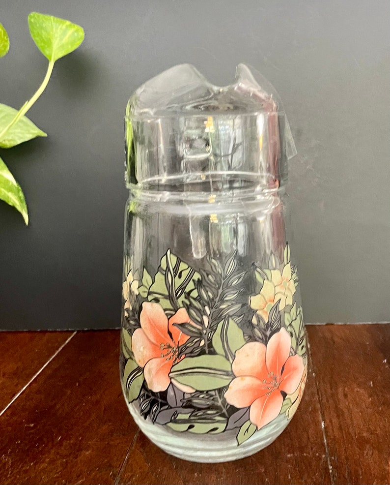 Puede incluir: Una jarra de vidrio transparente con un dise&ntilde;o floral. El dise&ntilde;o presenta flores rosas y amarillas con hojas verdes sobre un fondo negro.
