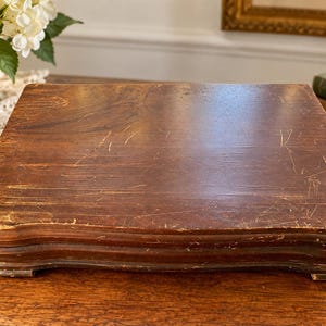 May include: A rectangular wooden display stand with a dark brown finish and visible surface wear, including scratches and scuffs. The stand features a tiered, molded edge design and rests on four short, integrated feet, providing a stable base.