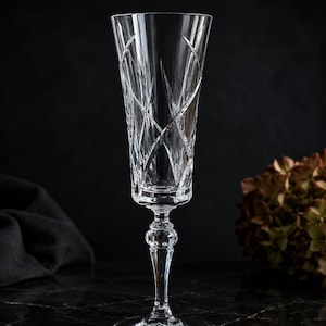 May include: A clear crystal champagne flute featuring a tall, slender bowl with deep, intersecting vertical and diagonal cut patterns. The stem includes a decorative faceted knob detail, leading down to a circular, flat, clear glass base.