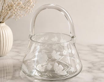 May include: A clear glass basket with a rounded handle and a wide, tapered base. The exterior surface of the glass features delicate, etched floral patterns, including various blossoms and leaves, creating a subtle, frosted white design.