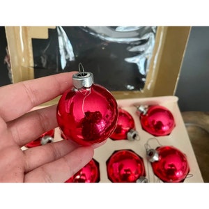 Puede incluir: Un conjunto de ocho adornos navide&ntilde;os de vidrio rojo con tapas plateadas. Los adornos est&aacute;n en una caja blanca con un fondo blanco.