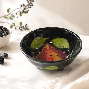 May include: A black ceramic berry bowl with a glossy finish, featuring a hand-painted red pear design with green leaves on the interior. The bowl has small drainage holes throughout the base and rests on three short, rounded feet for stability.