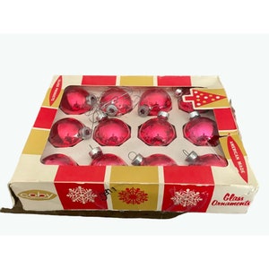 Puede incluir: Una caja de 12 adornos navide&ntilde;os de vidrio rosa. La caja es roja y dorada con un dise&ntilde;o de copo de nieve y las palabras "Coby Glass Ornaments American Made".