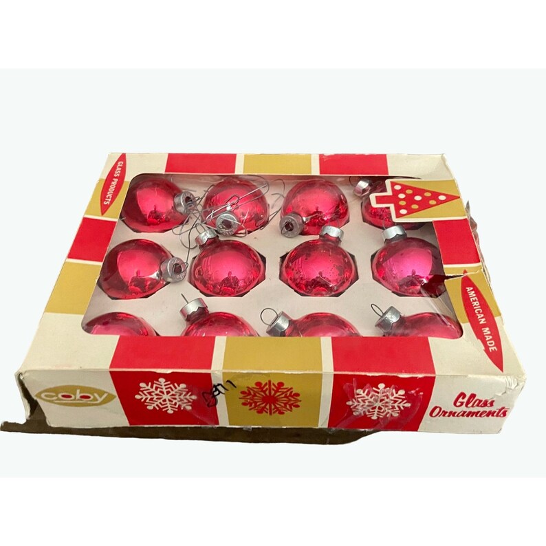 Puede incluir: Una caja de 12 adornos navide&ntilde;os de vidrio rosa. La caja es roja y dorada con un dise&ntilde;o de copo de nieve y las palabras "Coby Glass Ornaments American Made".