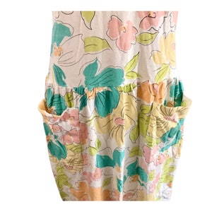 May include: A sleeveless dress with a floral pattern in pastel colors. The dress features large, stylized flowers in shades of pink, yellow, and teal, outlined in black. It has two large pockets and a gathered waistline, creating a relaxed, summery look.