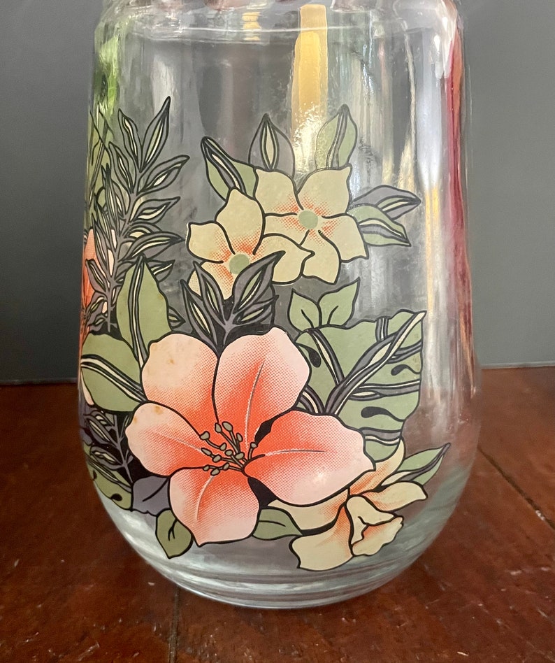 Puede incluir: Una jarra de vidrio transparente con un dise&ntilde;o floral. El dise&ntilde;o presenta flores rosas, amarillas y verdes con contornos negros.