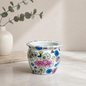 Könnte beinhalten: Ein weißer Keramikübertopf mit einem farbenfrohen, handbemalten Blumenmuster. Das Design zeigt Ansammlungen von rosa Pfingstrosen, kleinen gelben Gänseblümchen und blauen Blüten, die über die abgerundete Außenfläche des Topfes verteilt sind.