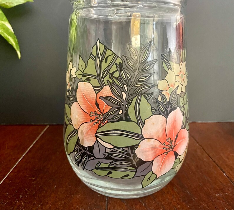 Puede incluir: Un tarro de vidrio transparente con un dise&ntilde;o floral. El dise&ntilde;o presenta flores rosas y naranjas con hojas verdes sobre un fondo negro.
