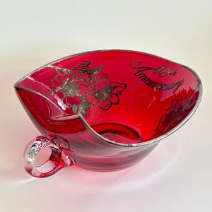 Puede incluir: Un cuenco de vidrio rojo rubí con borde y asa plateados. El cuenco presenta diseños florales plateados y el texto "40th Anniversary". El cuenco tiene una forma asimétrica única.
