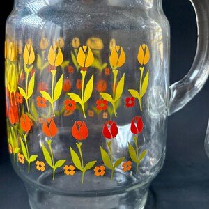 May include: A clear glass pitcher with a handle, featuring a repeating pattern of yellow, orange, and red tulips on a white background.