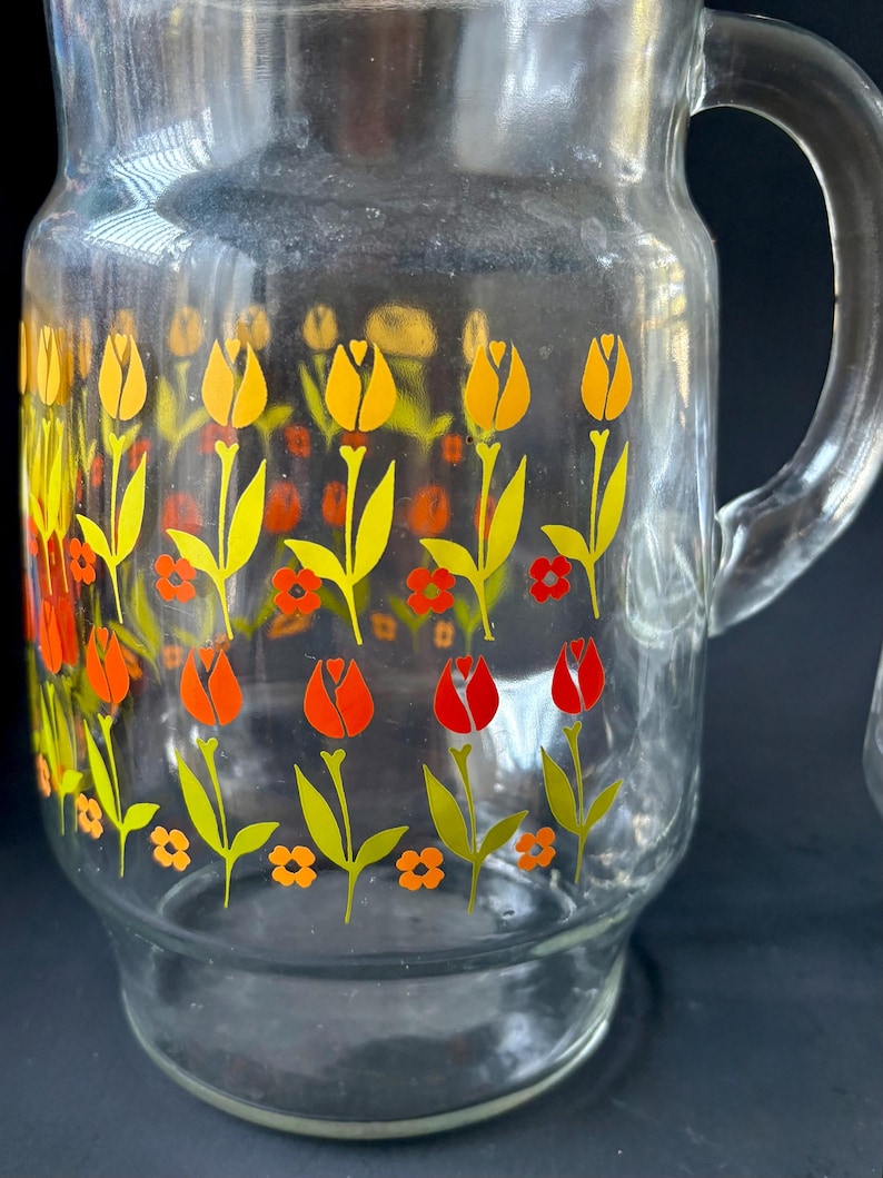 May include: A clear glass pitcher with a handle, featuring a repeating pattern of yellow, orange, and red tulips on a white background.