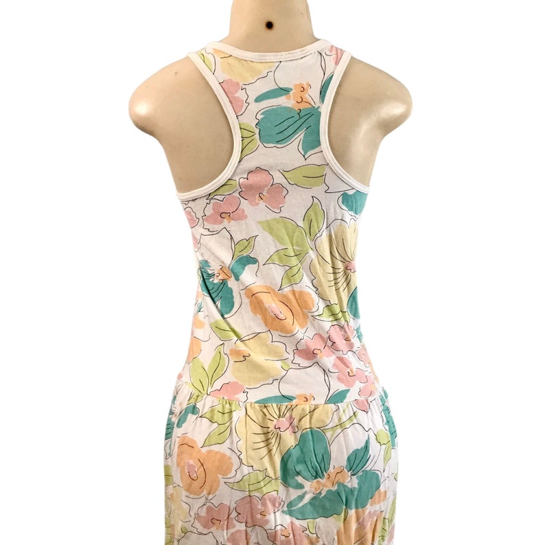 May include: White tank top dress with a floral pattern in pastel colors. The dress features a racerback design with a white trim. The floral print includes pink, yellow, and teal flowers with green leaves, creating a summery look.