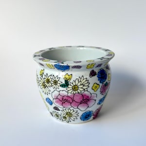 Op de afbeelding: Een kleine, ronde, witte keramische bloempot met een bloemenontwerp. De bloempot heeft een verscheidenheid aan kleurrijke bloemen, waaronder roze, gele en blauwe bloemen. De rand is versierd met kleine, kleurrijke bloemaccenten.