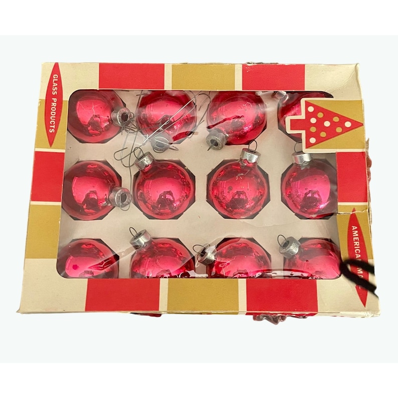 May include: A box of 12 red glass Christmas ornaments. The box is red and gold with the words 'Glass Products' and 'American Made' printed on it. The ornaments are in a clear plastic tray and are arranged in a grid pattern. Each ornament has a silver hanger.