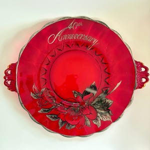 Puede incluir: Una fuente de vidrio rojo con borde y asas plateadas. La fuente presenta un diseño floral y el texto "40th Anniversary" en escritura plateada. La fuente tiene un borde festoneado.