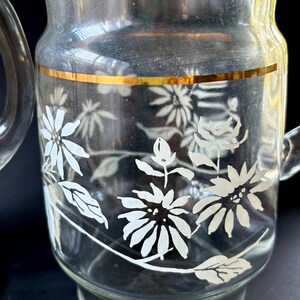 May include: A clear glass pitcher with a gold band around the top and a white floral design. The pitcher has a handle.