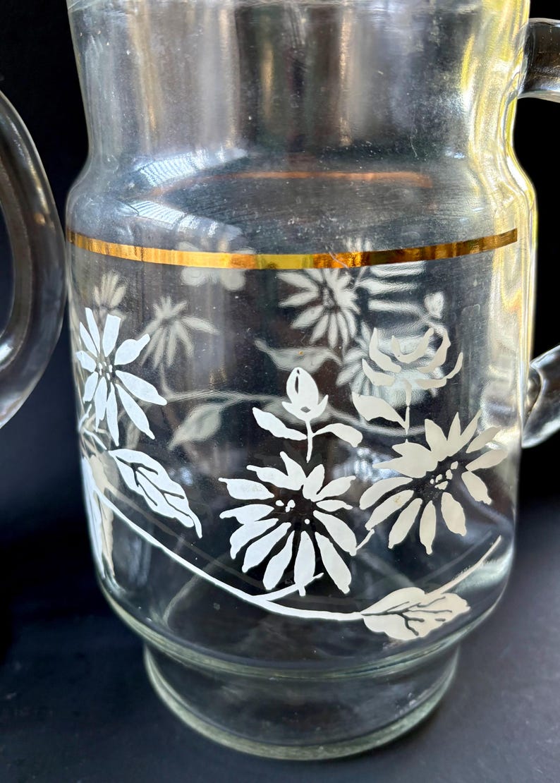 May include: A clear glass pitcher with a gold band around the top and a white floral design. The pitcher has a handle.