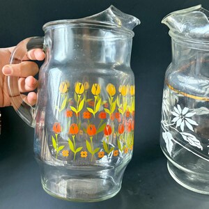 May include: Two glass pitchers with floral designs. One pitcher has a yellow, orange, and green floral pattern. The other pitcher has a white floral pattern with a gold band around the top.
