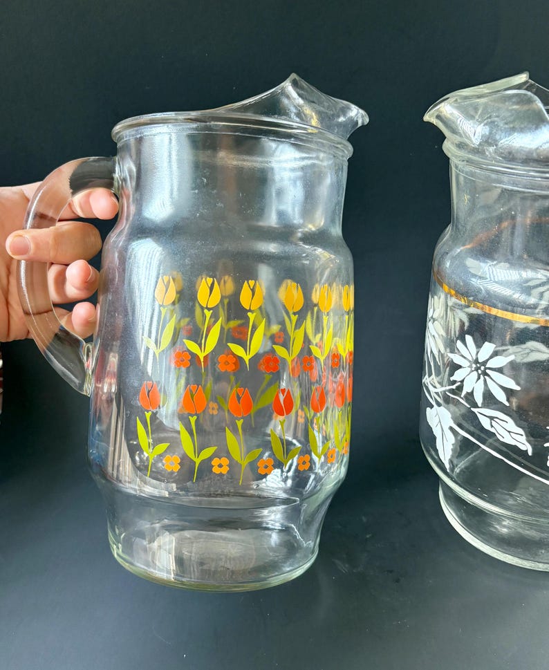 May include: Two glass pitchers with floral designs. One pitcher has a yellow, orange, and green floral pattern. The other pitcher has a white floral pattern with a gold band around the top.