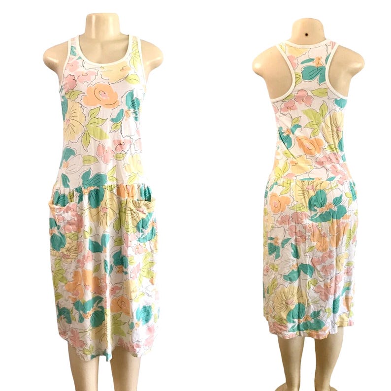 May include: A sleeveless floral dress with a white neckline and armholes. The dress features a pattern of large flowers in pastel colors, including pink, yellow, and teal, with two pockets at the waist. The dress is displayed on a mannequin.