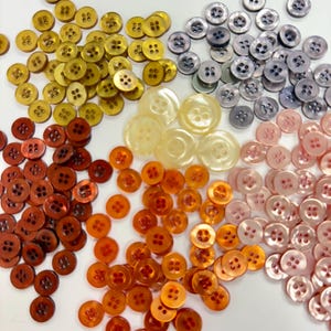 May include: A collection of various buttons in shades of gold, brown, orange, pink, and gray. The buttons are round with four holes and are arranged in separate piles. The buttons vary in size and have a glossy finish.