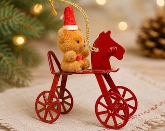May include: A small brown fuzzy teddy bear wearing a red and white Santa hat and a red bow tie, sitting on a red metal toy tricycle ornament. The ornament features a gold hanging string attached to the top of the bear's hat for display.