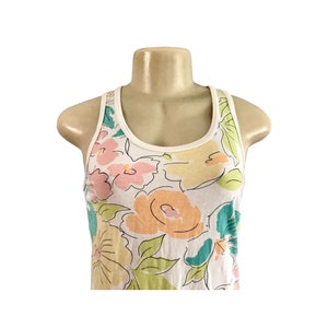 May include: White tank top with a floral pattern in pastel colors. The flowers are in shades of pink, yellow, orange, and teal, with green leaves. The tank top has a white trim around the neckline and armholes.