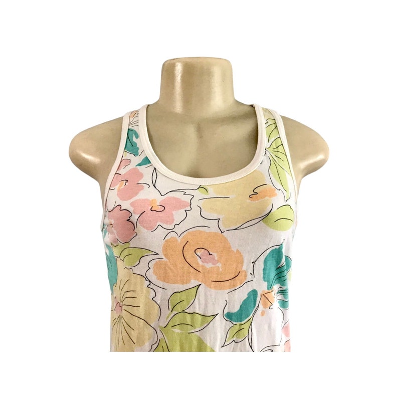 May include: White tank top with a floral pattern in pastel colors. The flowers are in shades of pink, yellow, orange, and teal, with green leaves. The tank top has a white trim around the neckline and armholes.