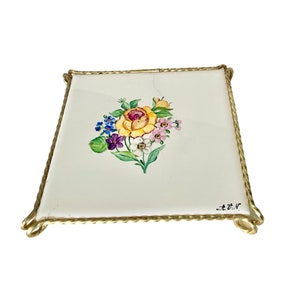 Puede incluir: Un azulejo de cerámica blanca con un diseño floral pintado de una rosa amarilla y otras flores en tonos de rosa, morado, azul y verde. El azulejo está enmarcado con un alambre de metal dorado y tiene la firma "Z.V.R." en tinta negra.