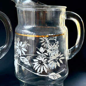 May include: A clear glass pitcher with a gold rim and white floral design. The pitcher has a handle and a wide mouth.