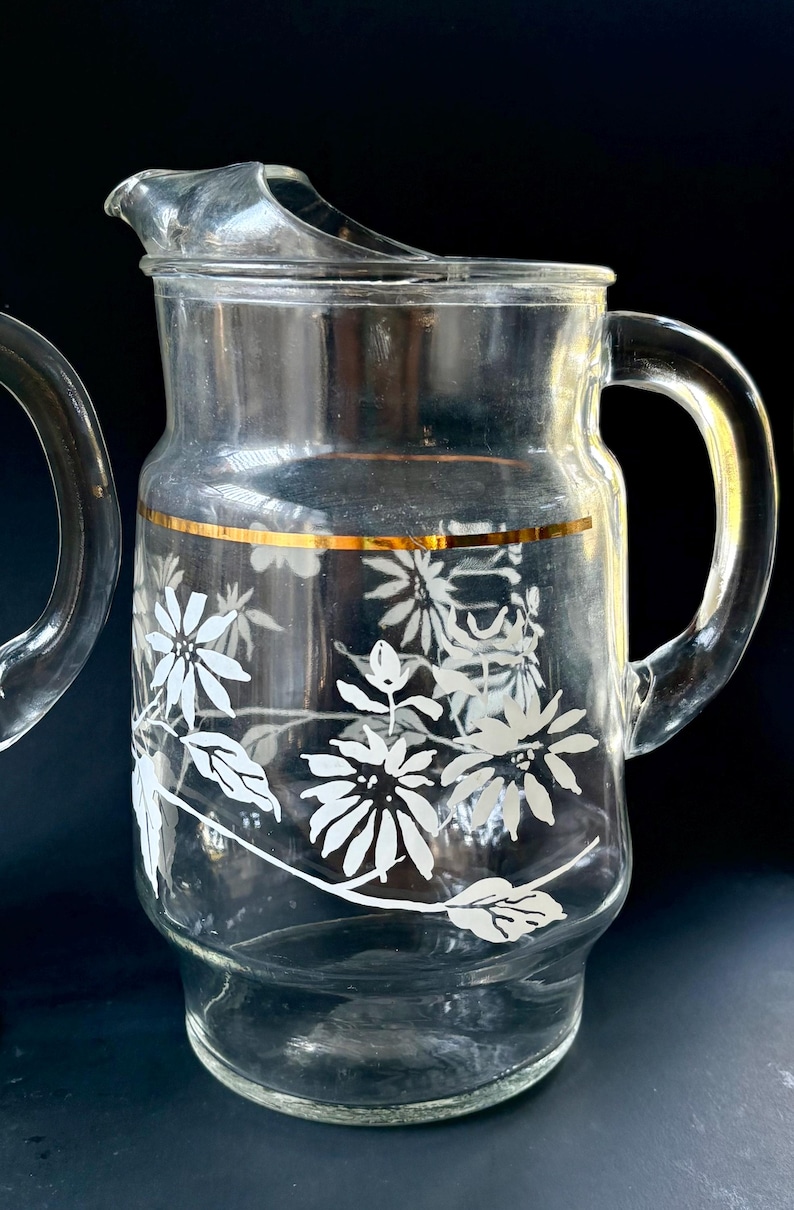 May include: A clear glass pitcher with a gold rim and white floral design. The pitcher has a handle and a wide mouth.