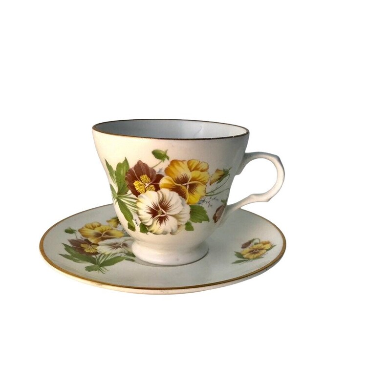 May include: A white teacup and saucer decorated with a floral pattern of yellow, brown, and white pansies. The cup and saucer have gold edging. A traditional tea set.