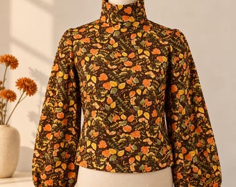 May include: A long-sleeved blouse with a high mock neck collar, featuring a dark brown base color with an all-over print of small autumn leaves in shades of orange, yellow, and green. The garment has full, gathered sleeves and a straight hem.