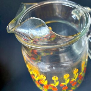 May include: A clear glass pitcher with a floral design. The pitcher has a yellow, orange, and red floral pattern around the bottom.