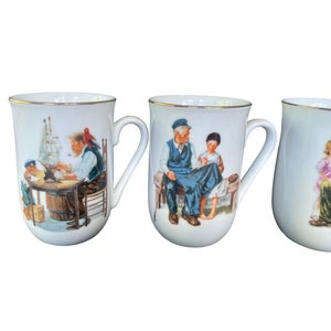 May include: Three white porcelain mugs with gold-trimmed rims, each featuring a detailed illustration. The first shows a person building a model ship, the second depicts an older person and a child sitting on a bench, and the third shows a child.