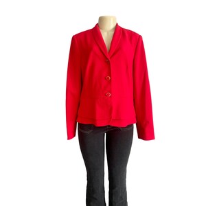 May include: A bright red blazer featuring a shawl collar, long sleeves, and three gold-toned buttons down the front. The jacket has a layered hemline detail and is displayed on a mannequin paired with dark charcoal gray denim jeans.